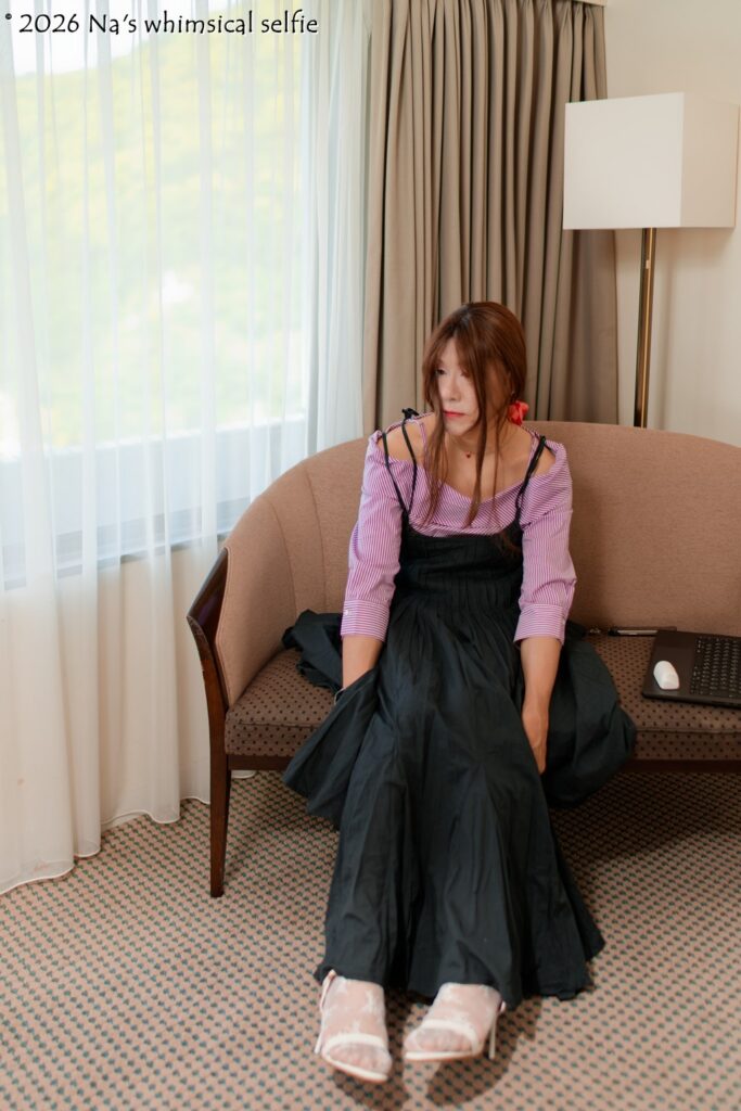 ピンクのオフショルダーブラウスと黒いロングスカートを着たNaが、ソファに浅く座って身体を前へ傾け、窓の方向をぼんやり見ている。